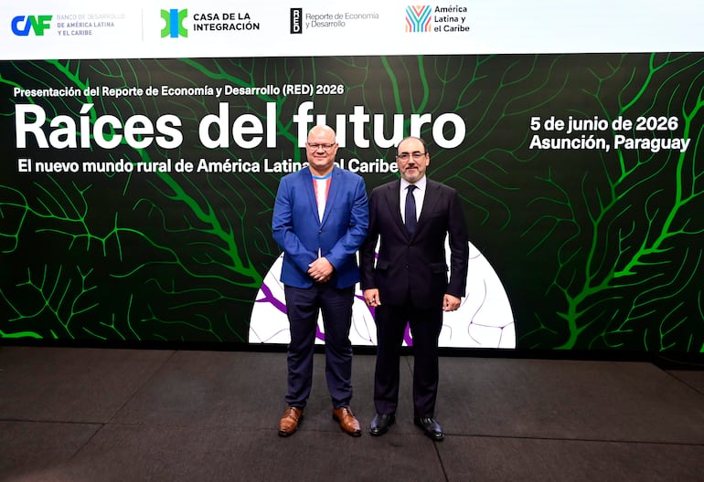 Óscar Lovera sonriente con saco azul, junto a otro hombre en traje negro en evento formal, fondo con gráfico de presentación.