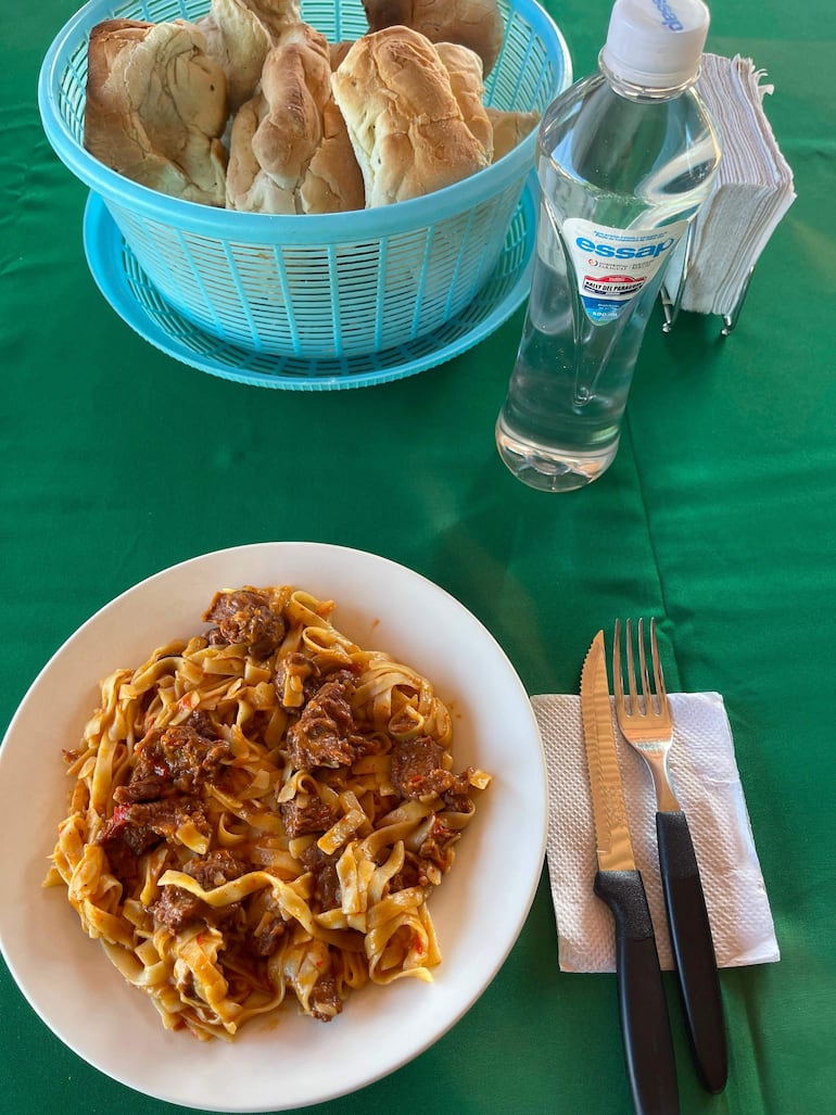 Mesa con mantel verde, platos de fideos y carne, canasta de pan y botella de agua Essap, sin personas visibles.