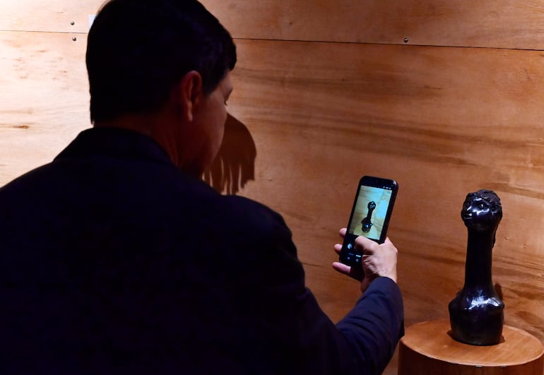 Hombre con traje oscuro de espaldas, usando un teléfono para fotografiar una escultura sobre pedestal de madera en un ambiente cultural.