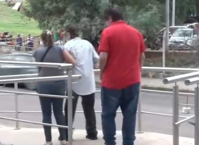 Mujer con camiseta gris y mochila ayuda a hombre en camiseta blanca, mientras otro hombre en camiseta roja observa.