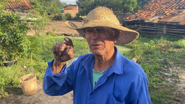 Hombre mayor con sombrero de paja y camisa azul, señalando con el dedo en un jardín rodeado de vegetación.