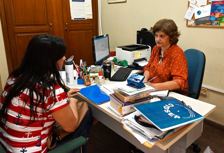 Dos mujeres, una de pie con camiseta de rayas, otra sentada en blusa naranja, conversan en una oficina con computadoras y documentos.