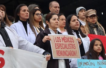 Mujer con gafas sostiene un cartel que dice '¡LOS MÉDICOS ESPECIALISTAS RENUNCIARON!', rodeada de manifestantes con batas blancas.