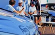 Cuatro personas en evento de automovilismo, dos hablando y escuchando, luciendo camisetas azules, con un coche de carreras visible al fondo.