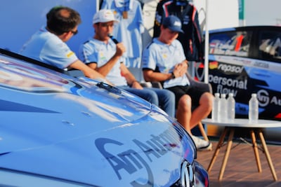 Cuatro personas en evento de automovilismo, dos hablando y escuchando, luciendo camisetas azules, con un coche de carreras visible al fondo.