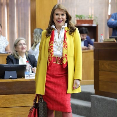 Celeste Amarilla sonríe en chaqueta amarilla y falda roja, interactuando con otra mujer en un ambiente formal.