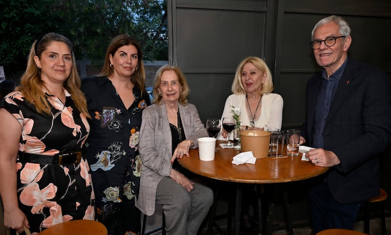 Daniel Nasta con gafas en un café, rodeado de tres mujeres sonriendo y una más participando, con copas de vino y una taza sobre la mesa.