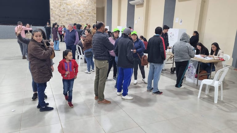 Mujer al frente levanta la mano, rodeada de adultos y un niño con camisa roja en un ambiente de ladrillo.