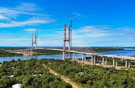 Puente atirantado en construcción sobre un río, rodeado de vegetación y cielo azul.