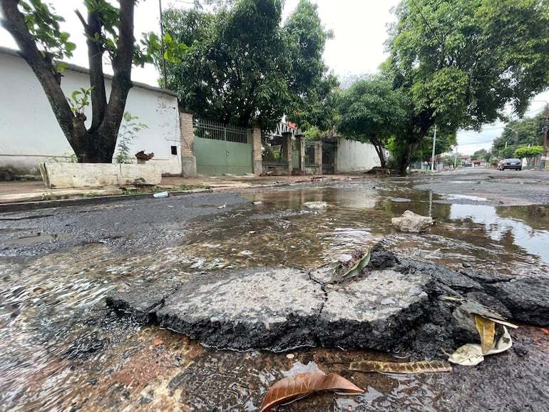 Calle en mal estado con baches y agua estancada, flanqueada por árboles y casas en el barrio Ybapobo.