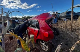 Una mujer se inclina dentro de un automóvil rojo dañado. Un hombre de pie observa, mientras otros dos hombres están en el fondo.