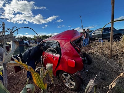 Una mujer se inclina dentro de un automóvil rojo dañado. Un hombre de pie observa, mientras otros dos hombres están en el fondo.