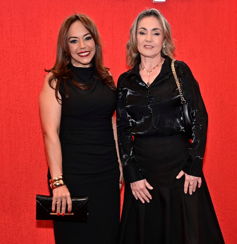 Dos mujeres de pie, una en vestido negro ajustado y otra en blusa y falda, sobre fondo rojo y sonrientes.