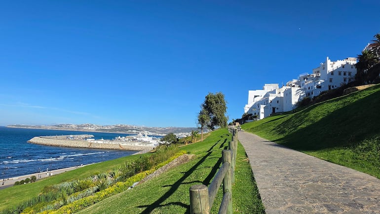 Hombre caminando por sendero verde junto a edificios blancos y un puerto con yates en un día soleado.