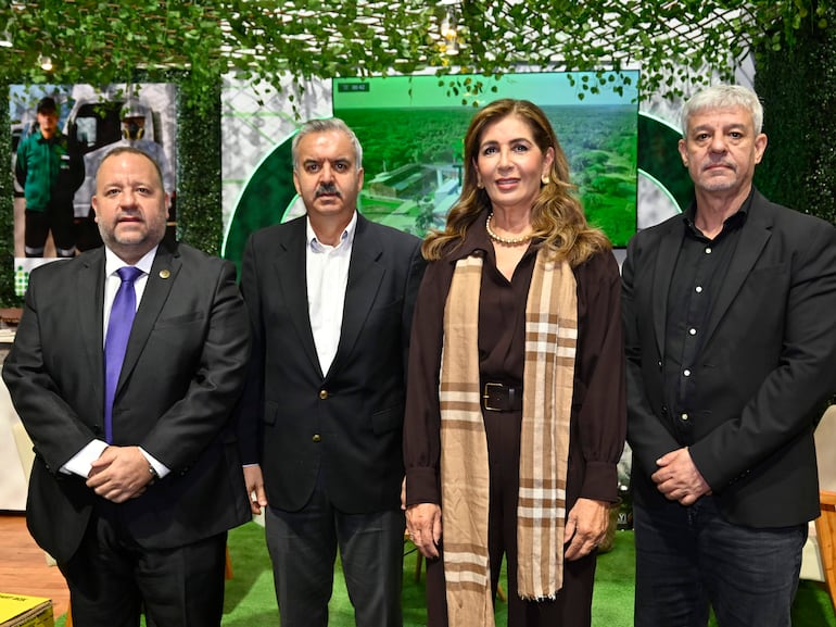 La mujer en blusa oscura y bufanda clara se destaca entre tres hombres con trajes oscuros, todos con expresiones serias en un ambiente formal.
