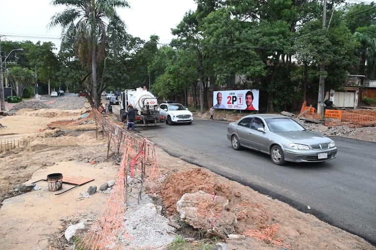 Calle en construcción con camión cisterna y vehículo blanco, rodeada de escombros y una barrera de seguridad.