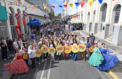 Mujer con blusa blanca y abrigo claro habla al micrófono, rodeada de personas con letreros amarillos en un evento al aire libre.