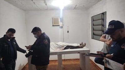 Cuerpo cubierto en mesa, cuatro hombres en ropa casual y uniforme examinan la escena en ambiente sombrío.