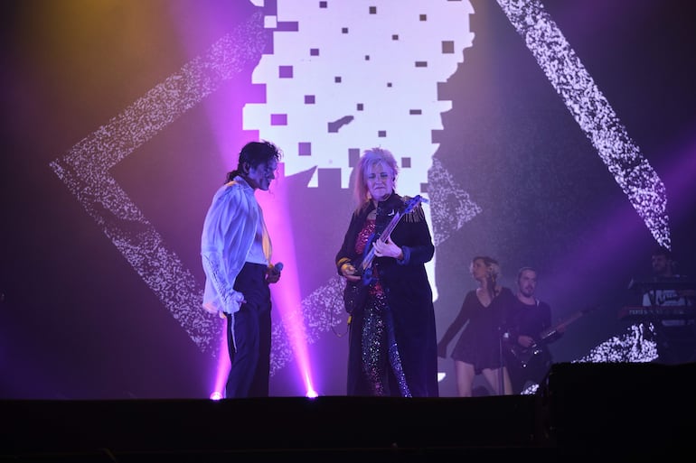 Lenny Jay con camisa blanca y Jennifer Batten con chaqueta oscura y guitarra, sobre un escenario iluminado con luces purpúreas.
