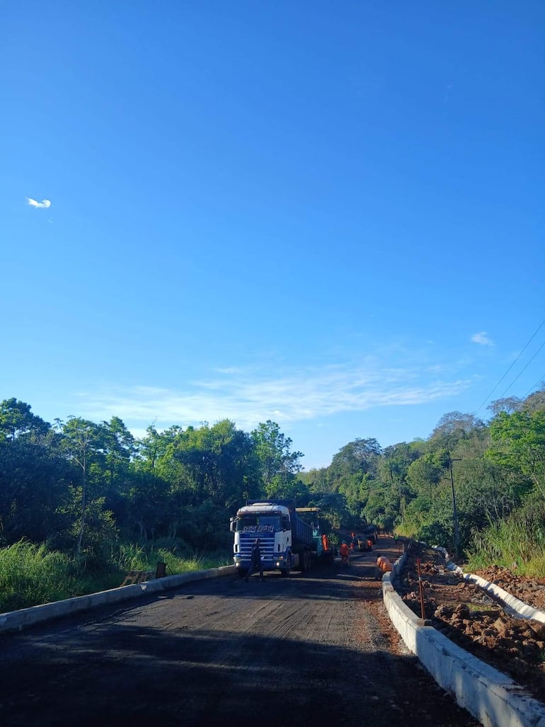 Camión volquete claro estacionado en la vía, trabajadores con chalecos de seguridad naranja operando maquinaria en un entorno rural.