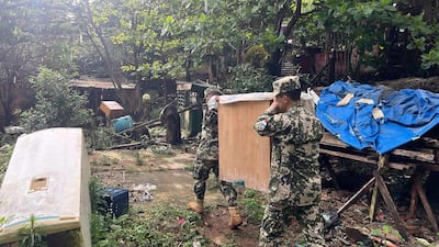 Dos soldados en uniforme camuflado levantan una plancha de madera en un entorno descuidado con vegetación y escombros.