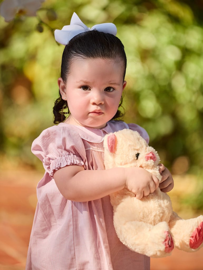 Niña pequeña con vestido rosa y lazo blanco, sosteniendo un muñeco de peluche en un entorno natural.