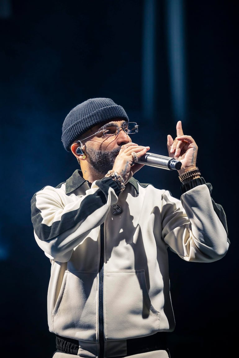 Arcángel, hombre con chaqueta clara y gorro de lana, canta con expresión enfocada en un escenario oscuro.