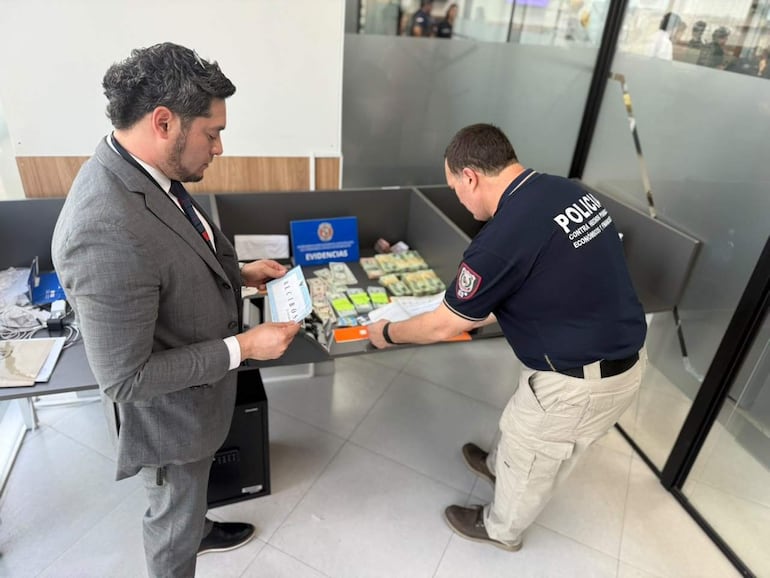Hombre con traje gris revisa documentos junto a un policía en camiseta. Mesa con billetes y documentos sobre fondo moderno.