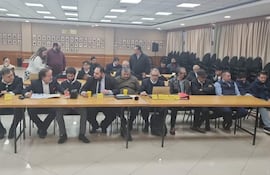Grupo de hombres de negocios y políticos en trajes formales discuten en mesa larga con micrófono, en un ambiente institucional.