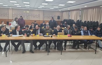 Grupo de hombres de negocios y políticos en trajes formales discuten en mesa larga con micrófono, en un ambiente institucional.