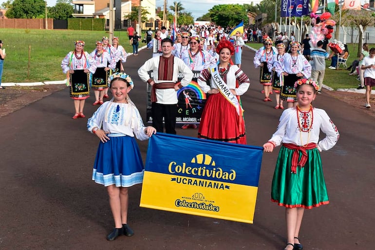 Niñas en trajes ucranianos desfilan con una bandera, sonriendo entre un grupo de adultos con atuendos tradicionales rojos y blancos.