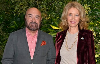 Hombre canoso con camisa rosa y saco gris, junto a mujer rubia con blusa gris y saco burdeos, en ambiente formal y decorado.
