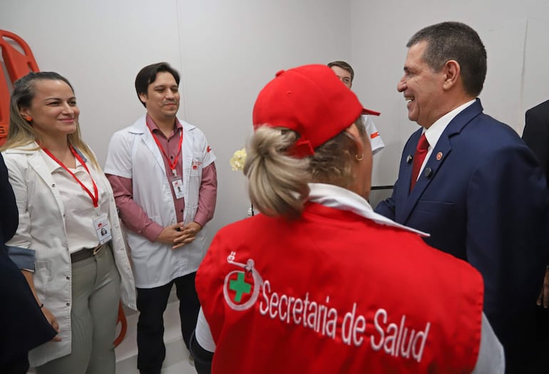 Hombre en traje oscuro sonríe y saluda a médicos en ambiente formal, con logo de la Secretaría de Salud visible.