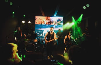La banda Ciudad Mansa en el escenario iluminado. Guitarrista sonriente al frente, bajista activa a la derecha y tecladista en el fondo.