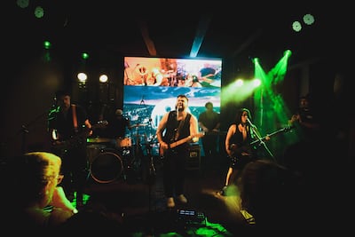 La banda Ciudad Mansa en el escenario iluminado. Guitarrista sonriente al frente, bajista activa a la derecha y tecladista en el fondo.