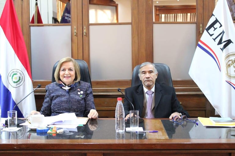 Dra. Alicia Pucheta vestida con chaqueta oscura y blusa clara, sonríe junto a Manuel Ramírez Candia en ambiente formal con banderas.