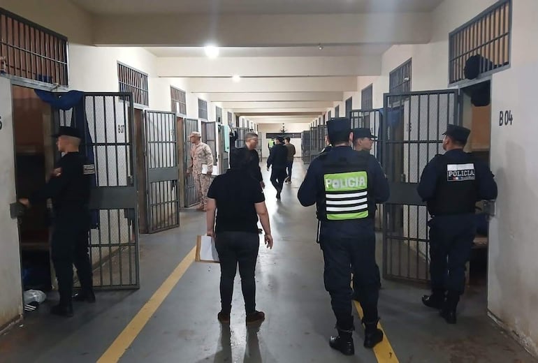 Oficiales de policía en uniforme revisan celdas en un ambiente penitenciario con puertas de rejas abiertas.