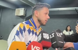 Ángel Romero viste chaqueta blanca y azul, rodeado de periodistas con micrófonos en un ambiente informal.