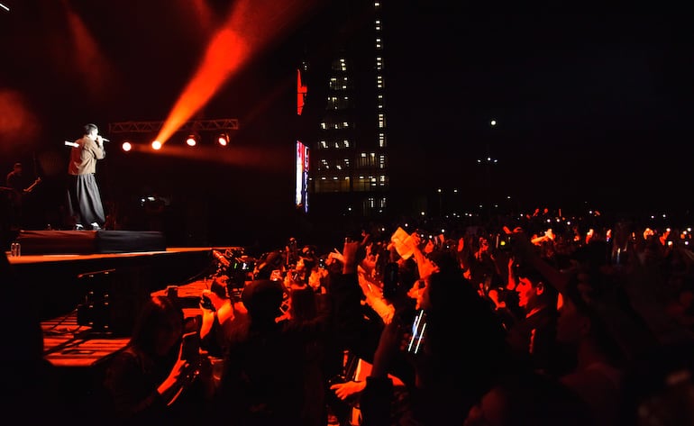 Milo J en escena con vestimenta oscura, animando a una multitud que levanta los teléfonos en un ambiente colorido.
