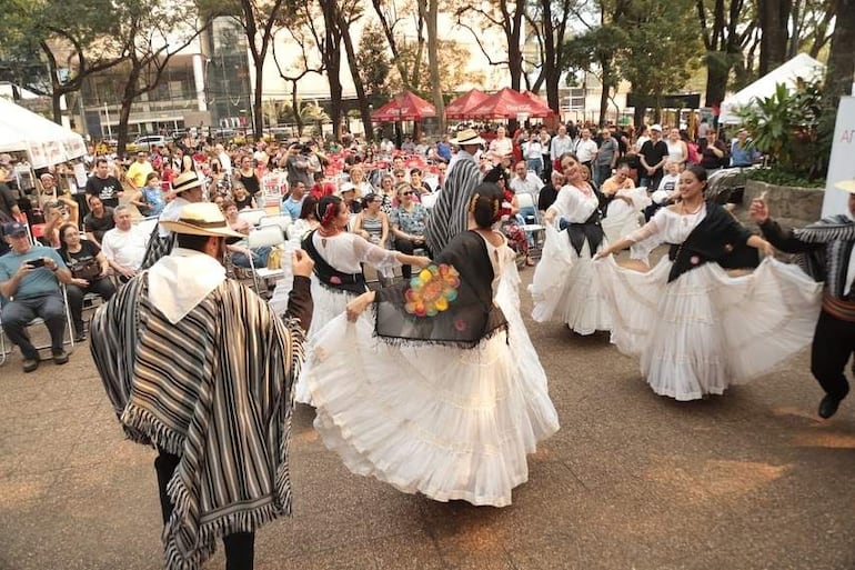 Bailarinas en trajes blancos con detalles negros y adornos florales, rodeadas de una audiencia diversa en un evento festivo.