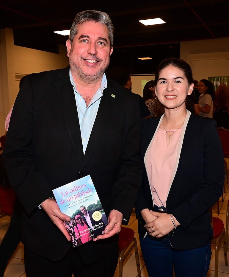 Hombre de traje negro sonriente sostiene 'La odisea de un jubilado', acompañado de mujer con blusa clara y blazer oscuro en sala de eventos.