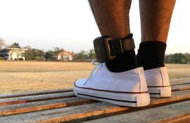 Un hombre parado en un espacio abierto al aire libre con championes blanco usa una tobillera electrónica una tobillera electrónica en el pie izquierdo.