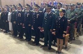 Grupo de personas en uniforme militar y trajes formales, con banderas de Paraguay de fondo, en un acto solemne.