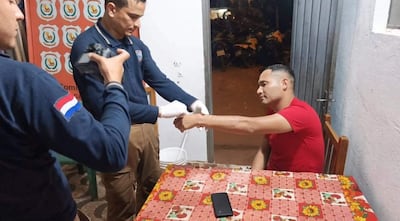 Hombre de camiseta roja sentado en mesa colorida, mientras dos hombres en pulóver oscuro atienden con vendaje.