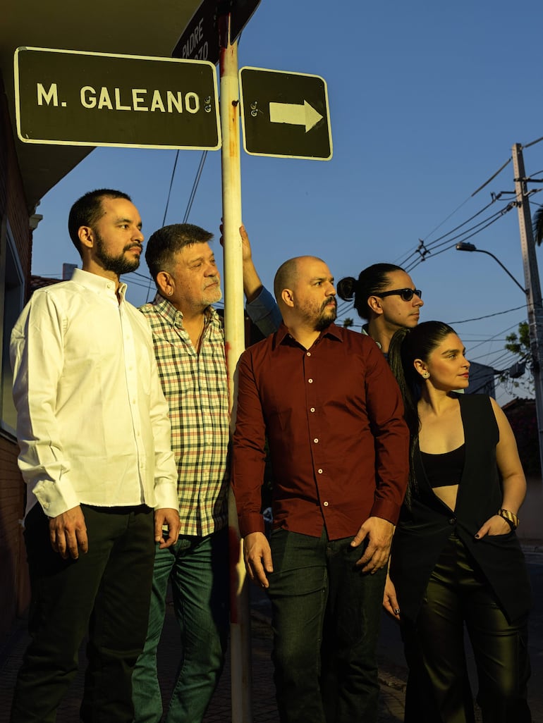 Cinco personas posan junto al cartel 'M. Galeano', vistiendo camisas coloridas y mostrando expresiones tranquilas al atardecer.