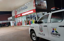 Estación de servicios iluminada, camioneta de policía visible y tres individuos en la escena; ambiente de tensión tras un intento de asalto.