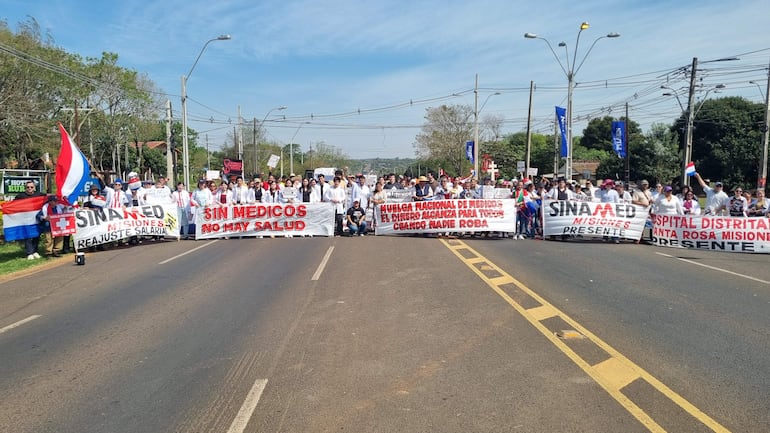 Grupo de médicos en batas blancas sosteniendo pancartas, algunos con instrumentos de protesta, en una vía pública entre árboles y edificios.