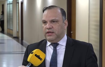Mario Varela con traje oscuro y corbata de puntos, habla ante micrófono en pasillo iluminado en un ambiente formal.
