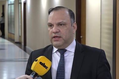 Mario Varela con traje oscuro y corbata de puntos, habla ante micrófono en pasillo iluminado en un ambiente formal.