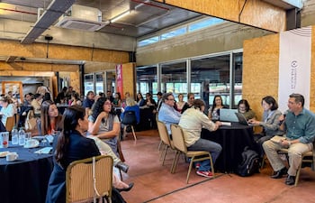 Grupo de cuatro personas en mesa, un hombre presenta con micrófono, mientras una mujer con laptop y otros escuchan en un ambiente profesional.